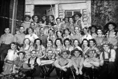 Photograph of forty two men and women dressed in costumes of gingham shirts,and cowboy hats in the case of the men, and spotted blouses and flowered dresses in the case of the women; they are posed in front of scenery in five rows; the photograph has been described as Nina Rosa, Murton