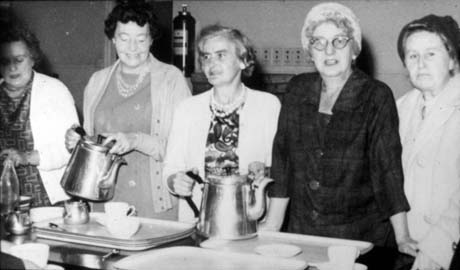 Photograph of five middle-aged women, wearing dresses and hats, standing with trays and cups in front of them; two of the women are holding large metal teapots; the photograph has been identified as Ladies Welfare Section - Mrs. Cocking, Murton