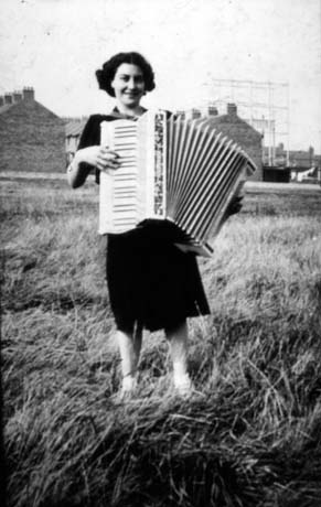 Photograph of a young woman, wearing a dark dress and holding an accordion, standing in a field with the ends of houses behind her; she has been identified as Rita Hall of Murton