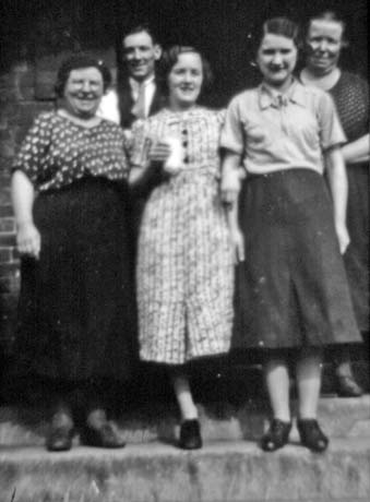 Photograph of three women, wearing skirts and blouses and a dress, standing on steps, with the head and shoulders of a man and a woman behind them; the woman in the dress is holding what appears to be a bottle of milk; they have been identified as Democratic Club Staff, Murton