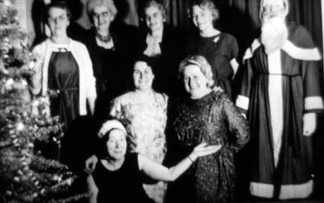 Photograph of eight women in dresses, posed in two rows, with an individual wearing a Father Christmas outfit; on the left of the picture is a Christmas tree; the photograph has been identified as Ladies Welfare Section Christmas Party, Murton