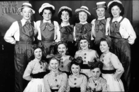 Photograph showing six young people dressed in boaters, blouses, bows and trousers with a bib, standing in a row with eight young people dressed in striped blouses, bows in their hair, dark sashes, and light skirts, kneeling in front of them; they have been described as Miss Daglish's Dance Troup - John Cocking, Murton