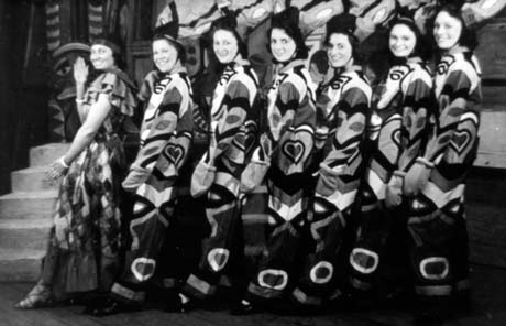 Photograph of seven women, with their left shoulders towards the camera, dancing and wearing loose, highly patterned trousers and blouses; they are dancing in front of scenery; they have bee described as Ladies' Chorus, Murton