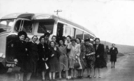 photograph of a group of eighteen women, wearing coats and dresses, standing outside a single-decker bus with a road and hills behind it; it appears to be raining and windy; the registration of the bus is HUP15..; they have been identified as members of Murton Womens' Institute on a trip