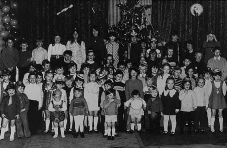 Photograph of a group of children, aged between three and thirteen years, wearing party hats, standing facing the camera; behind them two Christmas trees can be seen indistinctly; the photograph has been identified as Royal Order of Buffaloes Children's Christmas Party in Murton
