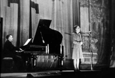 Photograph showing a woman in a suit standing at the edge of a stage with a man sitting at a grand piano accompanying her; there is a highly decorated pilaster at the side of the stage and a high curtain behind them; the photograph has been identified as Pianist and Singer - Theatre Royal, Seaham