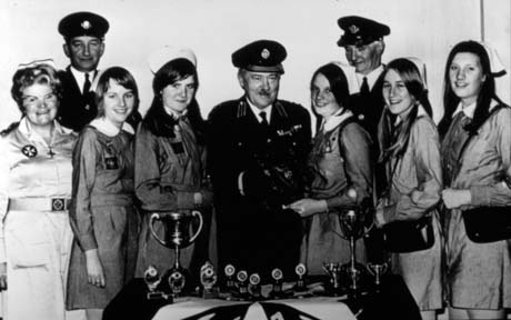Photograph showing five girls, aged approximately fourteen years, wearing a uniform, standing on either side of a man wearing a uniform; on the left is a woman in a uniform and two men in uniform are standing behind the others in the group; in front of them are four trophy cups and eight small trophies; they have been identified as St. John Ambulance Cadets Presentation - Mr. and Mrs. S. Cummings and Dr. Haydock