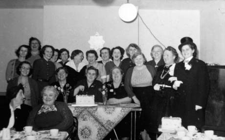Photograph of sixteen women, wearing dresses and skirts, standing against the wall of a room; in front of them are three women sitting at a table with an iced celebration cake on it; in front of them are two women sitting at another table with cups and plates on it; a woman wearing a dinner jacket and top hat is standing on the right of the group standing at the back; they are described as The British Legion Committee in the Old Hut at the bottom of the terrace, Murton