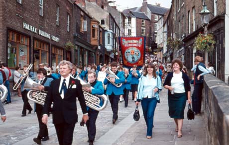 Photograph showing the members of a band walking down Elvet Bridge in Durham City towards the camera, in front of a miners' banner, which is unidentifiable; the buildings on either side and onlookers can be seen