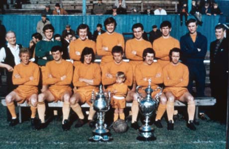 Photograph of eleven men in orange football strip, posed on a bench with the stands of a stadium and a small number of spectators behind them; they are accompanied by four men and a small boy, aged approximately two years, standing in front of them in the strip; also in front of them is a football and two trophy cups; they have been identified as Murton Colliery Welfare Athletic Football Club
