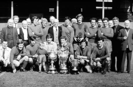 Photograph showing eleven men in football strip posed with twelve other men in a football stadium; in front of the group are three trophy cups and a small boy aged approximately two years; the photograph has been described as Colliery Football Team - Shipowners' Cup, Monkwearmouth League, Murton