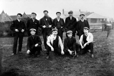 Photograph of six men, in suits and caps, standing in a row behind four other men, who are squatting on the ground; two men have cloths over their shoulders and one is holding an animal; behind them are hayricks and farm buildings; they have been identified as being in Murton