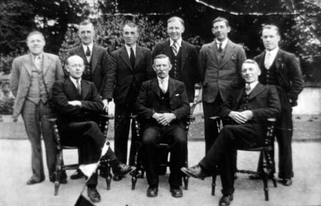Photograph of six men in suits and ties standing behind three men who are sitting in Windsor chairs; behind the men are a path and bushes; they have been described as being at Conishead Priory Convalescent Home in Cumberland