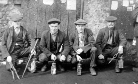Photograph of four men crouching on the ground in front of a rough wall and door; they are wearing jackets, waistcoats, boots and caps; each is holding a miner's lamp and those on either end are holding a pointed metal instrument with an eyelet in its middle; they have been described as Miners at Colliery with Lamps, Murton