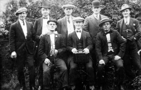 Photograph showing eight men posed in front of trees; the man at the left on the front row has a whitened face, a bow tie and a cane in his hand; next to him is a man with a cap. a bow tie and a small suitcase; next to him is a man with a boater and a blackened face; one man on the back row is wearing a top hat and bow tie and another has a bowler hat; they have been identified as The MacDuds Concert Party - James Campbell, Edgar White, Dobbs, Chapman, Mr. Daglish, Diddler Curtis, Brian Foster
