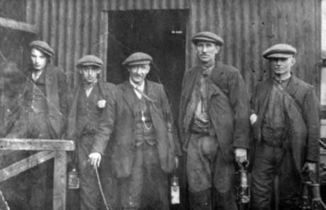 Photograph of five men standing in front of a large corrugated iron building; they are dressed in jackets, waistcoats, scarves and caps; they are carrying miner's lamps; the photograph has been described as Derek Gibson - Miners, Murton