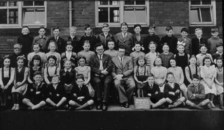 Photograph of forty one children, aged approximately nine years, posed in three rows against the wall of a brick building; they have been described as pupils at a school in Murton; they are accompanied by two men on the front row