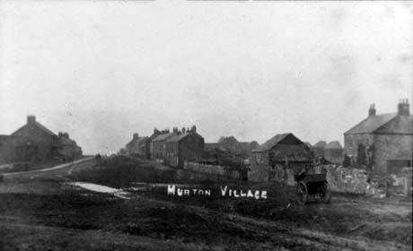 Postcard photograph entitled Murton Village, showing two rows of houses on either side of a green; in the foreground on the right of the photograph are the buildings of what appears to be a farm; a cart is in front of the wall round the farm