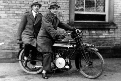 Photograph of two young men wearing overcoats and trilby hats sitting on a motor cycle with the wall and window of a house behind them; the registration of the motor cycle is as follows: HN218; the men have been identified as Billie Olaman and David Lowery