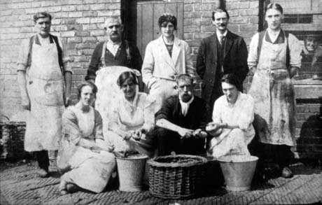 Photograph showing three men wearing aprons, a woman in an overall, and a man in a suit, standing against a brick wall with a door and part of a window in view; a man's face can be seen in the window; in front of the four against the wall, are three women and one man crouching on the ground, with a basket and two buckets in front of them; one of the women and the man are peeling potatoes; the photograph has been identified as Waterworks Soup Kitchen, Murton