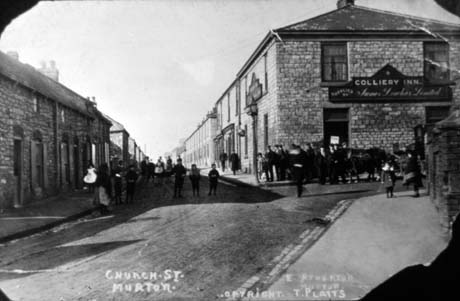 Postcard photograph entitled Church St. Murton. Copyright T. Platts. E. Atherton., showing a road with terraced houses on either side receding from the camera; on the end of the road on the right is the end and front of the Colliery Inn; groups of people can be seen indistinctly in the road and on the pavement