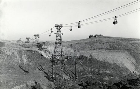 Photograph showing pylons in a line across country supporting cables on which there are containers of coal waste passing to and fro; a pit heap with a machine on it is on the right of the picture; the photograph has ben identified as being in Murton