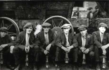 Photograph of five men and a boy, in jackets, caps and shirts, holding miners' lamps ,squatting in front of the side of a coal waggon, the wheels of which can be seen in detail; indistinct heads of three men looking between the waggons, can be seen; the men have been identified as being in Murton