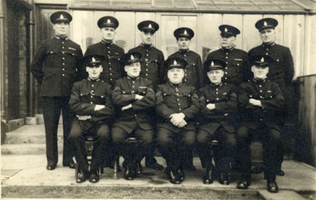 Photograph of six men in uniform standing in a row in front of low building of metal or glass; five men, also in uniform, are sitting in front of them; they have been identified as Special Constables in Murton during the Second World War