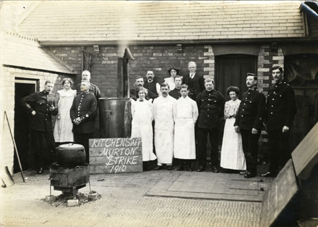 Photograph showing a yard with a single-storey building along the back and left side; against the back building, four women in long ankle-length aprons, a man in a long apron, two men in suits and eight policemen, are standing; a boiler with a chimney and a pot on a brazier can also be seen; in front of the boiler is a notice reading; Kitchen Staff Murton Strike 1910