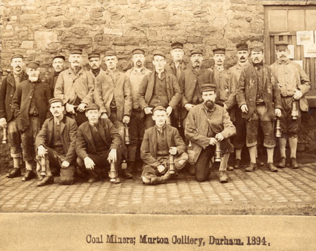 Photograph with the caption: Coal Miners; Murton Colliery, Durham. 1894., showing fourteen men wearing caps, jackets, waistcoats, scarves, breeches and boots, standing against a rough stone wall; in front of them, four men, dressed in the same way as those standing, are squatting on the ground; all the men are carrying miners' lamps