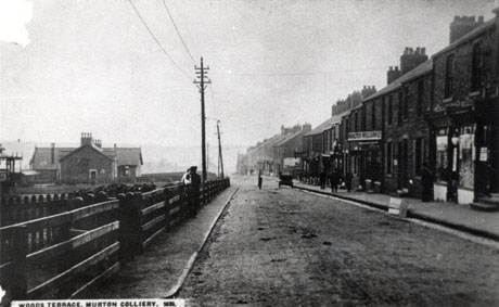 Postcard photograph entitled Woods Terrace, Murton Colliery. 1636, showing a road running away from the camera with a fence on the left and two houses in the distance behind the fence; on the right of the photograph is a line of shops and terraced houses, with a sign advertising Walter Willson the only one discernable; a car is parked with its back to the camera in the middle distance; indistinct people can be seen on the pavement and leaning over the fence