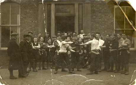 Photograph showing a group of men in a semi-circle standing watching two men stripped to the waist, sparring with boxing gloves on; behind the men is the facade of a large brick house with long sash windows and a high door with pillars either side of it; the men are wearing gaiters, breeches and shirts and overcoats; they have been identified as the Murton Battalion of the Territorial Army in 1900