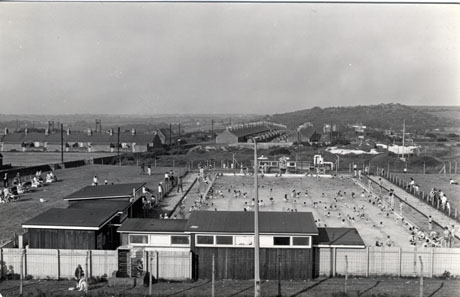 Photograph, taken from above, of a swimming pool with rows of terraced houses. hills and the pylons of the aerial ropeway, in the distance; the pool has a fence round it and low buildings on the left, presumably changing rooms; the pool is full of swimmers; the pool has been identified as being in Murton