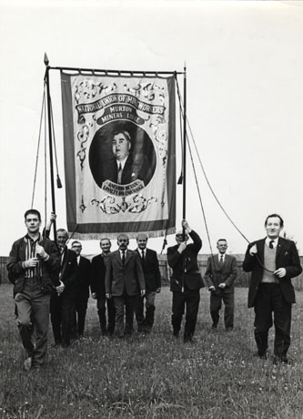 Photograph showing nine men, all but one wearing suits, walking with the Murton Lodge banner in a field; the banner carries a portrait of Aneurin Bevan, minister in the government of Clement Attlee, 1945- 1950, and founder of the National Health Service; under his portrait are the words: Aneurin Bevan Loyalty and Endurance