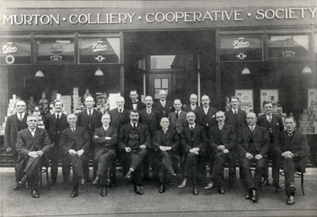 Photograph showing thirteen men standing in front of the windows of Murton Colliery Cooperative Society; in front of them are ten men sitting on chairs; all the men are wearing suits and ties; piles of tins and packets can be seen in the windows