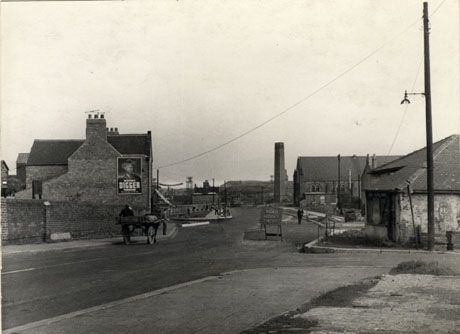 Photograph showing a road running away from the camera with a pit heap and aerial line for the conveyance of coal waste in the distance; on the right of the photograph a large chimney and the side of a chapel can be seen; a horse and cart and a sign warning of road works can be seen; a low dilapidated building can be seen on the right of the picture, which has been identified as being in Murton