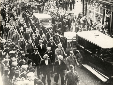 Photograph showing a procession of cars pulling unidentifiable objects with men in uniform and men in civilian dress walking along beside the cars; some of the men are holding what appear to be rifles; crowds are lining the route of the procession; a shop with T. Hanley and Fresh Farm Eggs on its front, can be seen; the photograph has been identified as being in Murton