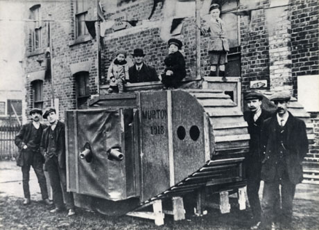 Photograph showing a mock tank with caterpillar tracks and the barrels of two guns sticking out its side and the words Murton 1918 written on it; four men, wearing suits and caps, are standing near it and a man, wearing a suit, overcoat and bowler hat and three children are standing on it; behind the tank is a brick building with a plaque reading: Miners Hall on it; there is a string of bunting above the tank and it is possible that the photograph is recording celebrations to mark the end of the First World War