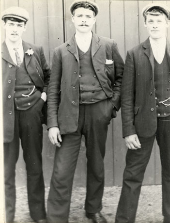 Photograph of three young men standing in a row against a wooden wall or fence; they are wearing suits, waistcoats, ties and caps; one of them has a flower in his button hole