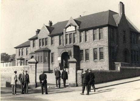 Photograph showing the front and side of a large brick building with bay windows and a large porch with steps leading to it; there is yard and wall round the building which bears a sign reading: Murton National Democratic Social Club Ltd.; eight men are entering the yard and two are standing at the top of the steps to the building