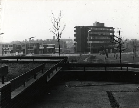 Photograph of the exterior of Brandlings Court, Peterlee, showing a large block in the distance on the right and a lower block, with shop fronts, on the left in the distance; in the foreground are low fences and walls