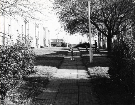 Photograph showing a path running up-hill away from the camera; on either side of the path is grass and, beyond the grass, the exterior of linked buildings; a small girl and a man and a small boy can be seen in the distance on the path and the grass