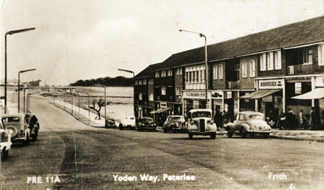 Postcard photograph entitled PRE 11A Yoden way, Peterlee. Frith, showing a parade of shops on the right of the road; the shops include Broughs and Woolworths; no others can be identified; beyond the shops are open fields; eight cars can be seen in the road, including one with the registration DUP 174; indistinct figures can be seen in front of the shops