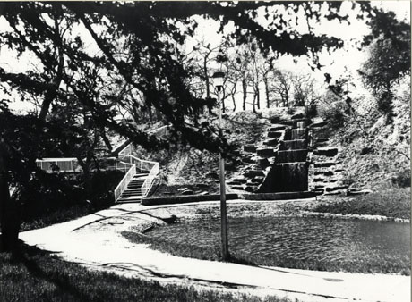 Photograph of a cascade of water coming down a slope; on the left of the cascade is a flight of steps coming down the slope; in front of the slope is a pond, a footpath round the pond and a street light; the photograph has been described as Waterfall, Back of Norseman Hotel, Peterlee