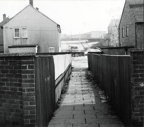 Photograph of a view between the fences of two conventionally built houses, looking towards a slope with, at the top in the distance, low buildings, possibly factory units; the photograph has been identified as View From Yoden Road, Peterlee