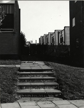 Photograph showing a fight of five brick steps in a grass bank leading to a fence behind which linked buildings of two storeys can be seen;the photograph has been identified as Manor Way, Peterlee, 1976