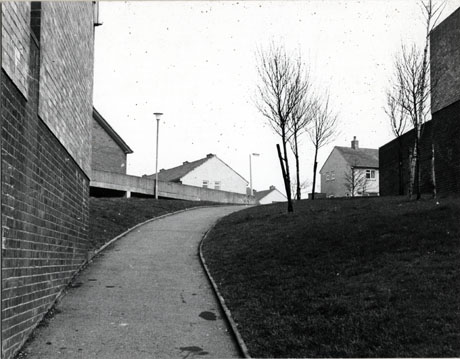 Photograph of a path running uphill away from the camera towards the right; a brick wall is in the left of the picture and an expanse of grass on the right; in the centre, in the distance, are the tops of houses with ridge roofs; the photograph has been identified as Brandlings Court, Peterlee