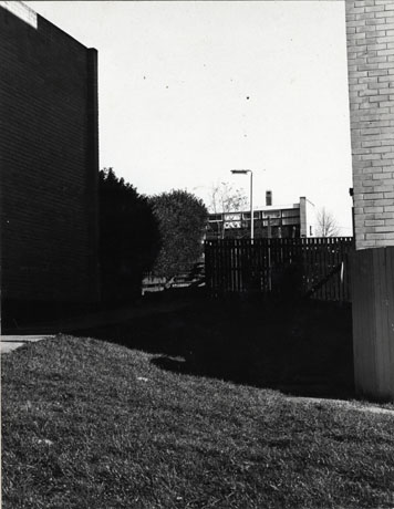 Photograph, taken between two buildings, of a two-storey building in the distance, behind a fence, bushes and a lamp post; the photograph has been identified as Manor Way, Peterlee, 1976