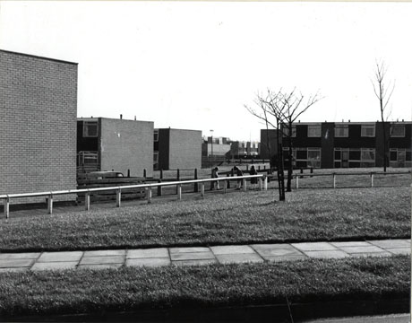 Photograph of an expanse of grass surrounded with alow fence; beyond the fence are three children standing on a path; beyond the children are the blank brick ends of two-storey houses; in the distance, on the right of the picture the facades of the two-storey houses can be seen; the houses have been identified as being in Peterlee