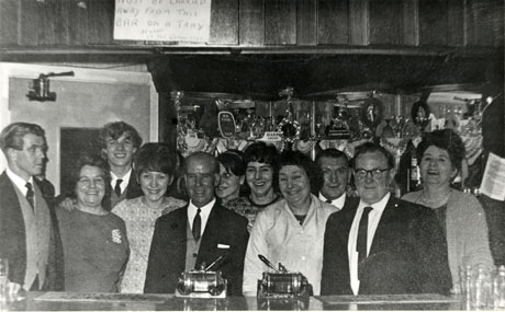 Photograph of five men and six women standing behind the bar of the working men's club in Peterlee; they have been identified as the Turton family; behind the group, shelves containing glasses, and, in front of the group, the surface of the bar, can be seen